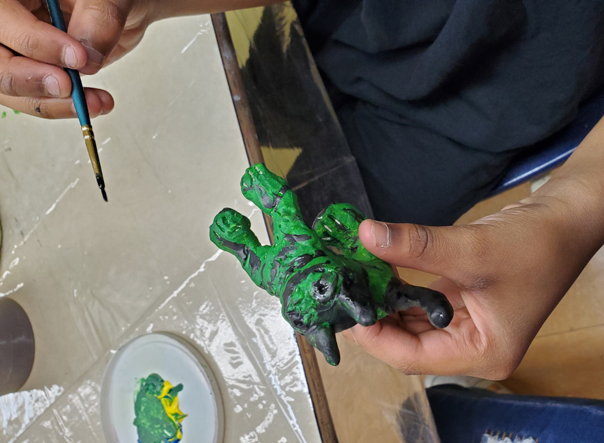 mains d'un élève en train de peindre en vert et noir une figurine d'animal inventé de forme féline