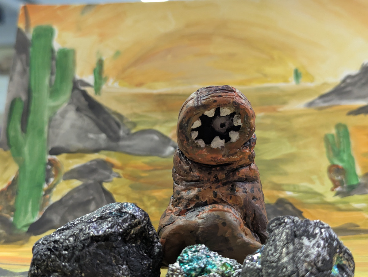 Sculpture d’une créature extra-terrestre, sur un fond peint représentant des rochers, des cactus et du sable