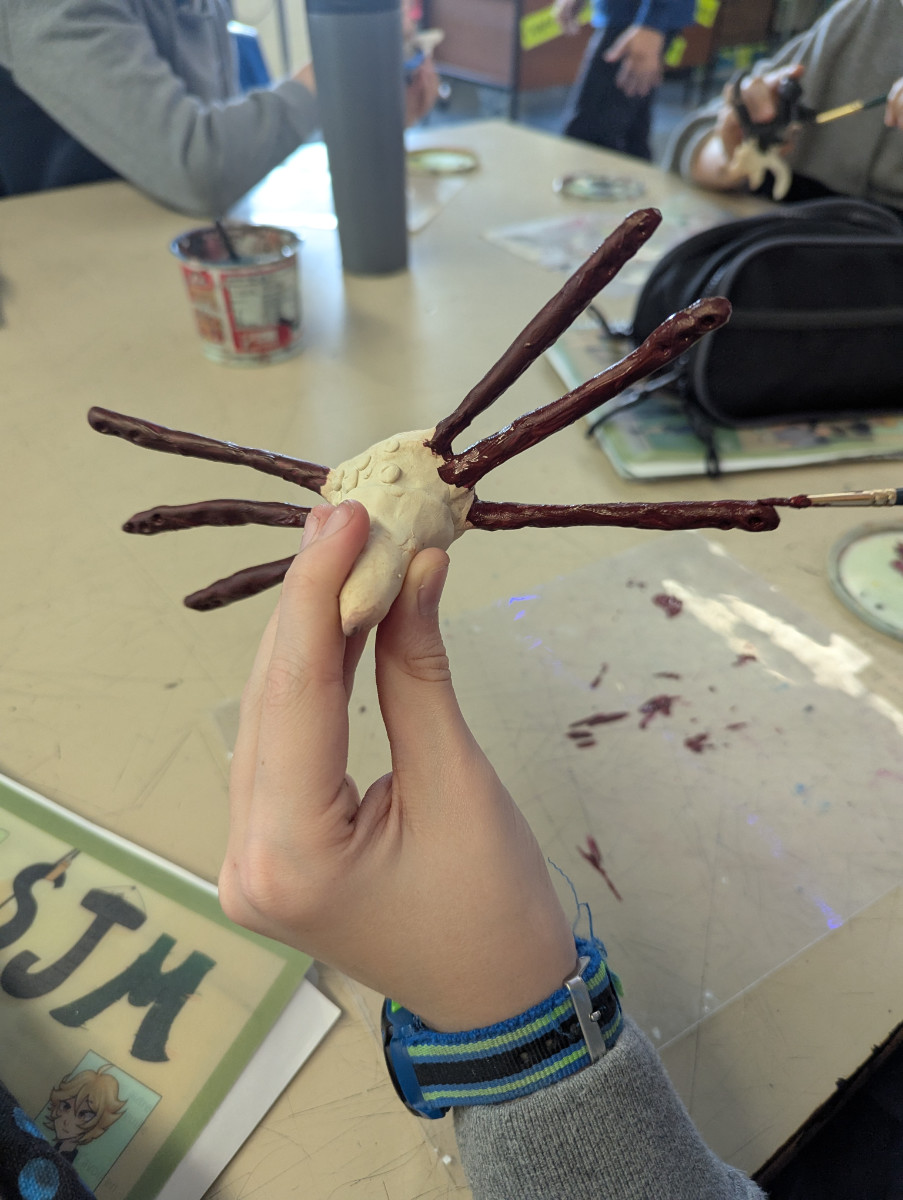 main d'élève peignant une figurine d'argile polymère. En fond, plusieurs autres élèves peignent aussi leur projet