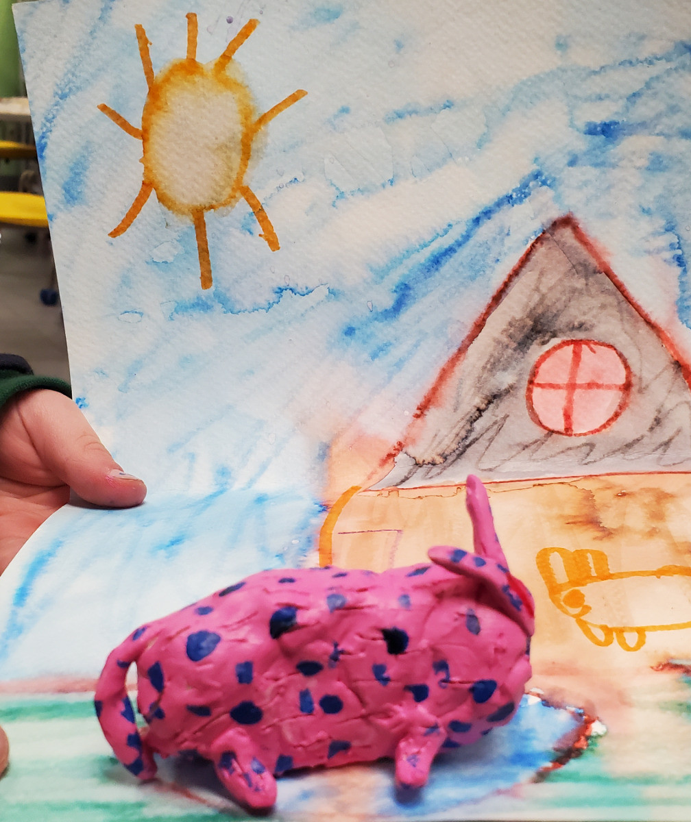 Figurine d’animal inventé rose à pois sur un dessin où il y a une maison, un soleil et un ciel bleu.