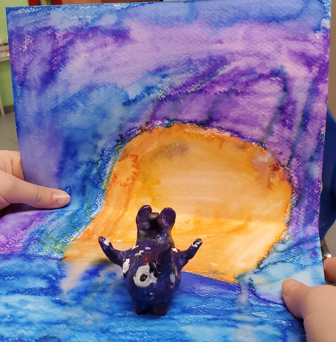 Figurine d’animal inventé peinte sur un arrière plan de style aquarelle. Les couleurs principales de l'oeuvre sont le jaune, le bleu et le violet.