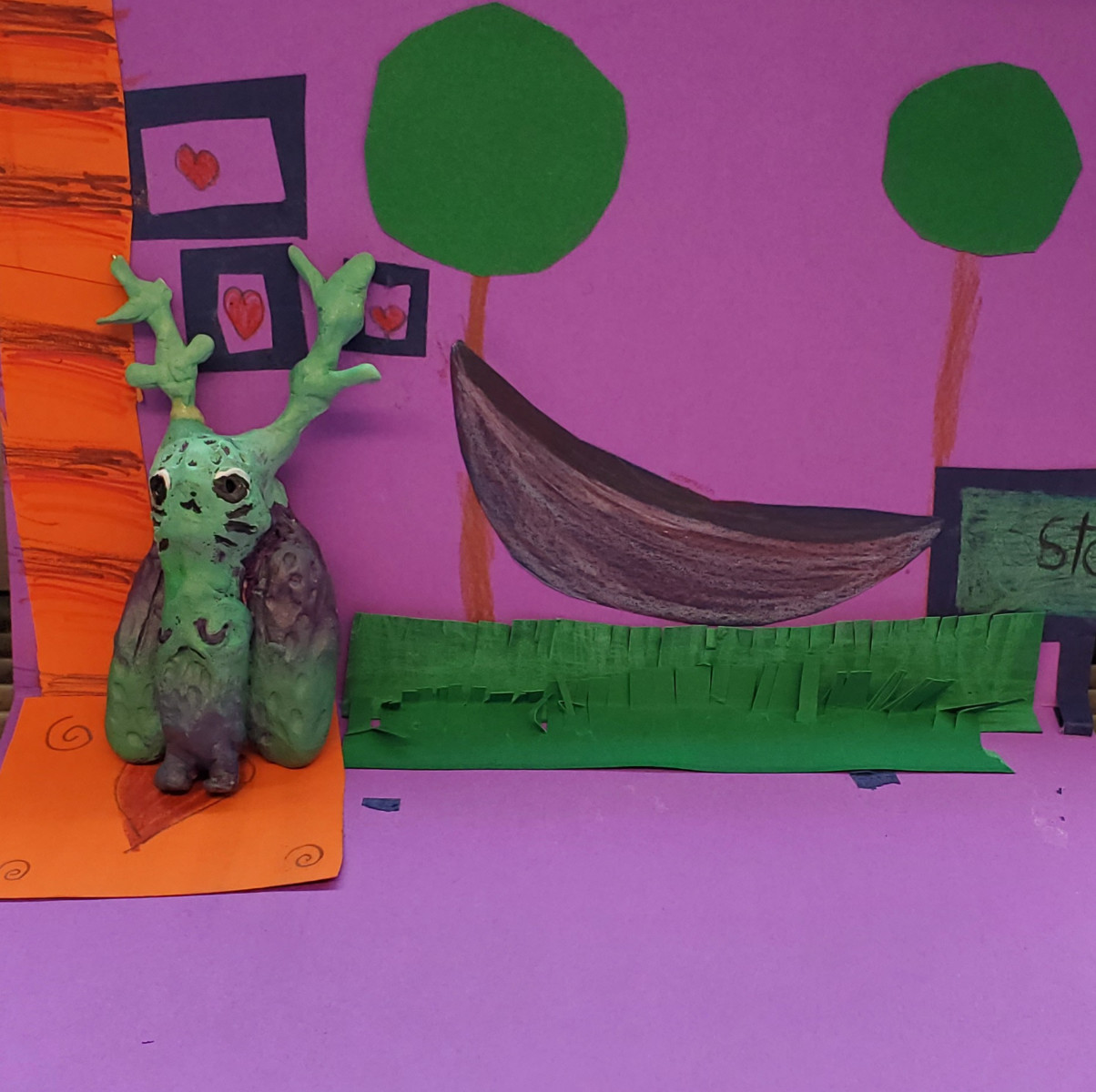 Figurine d’animal inventé peinte dans des tons de turquoise et de violet. À l'arrière-plan, de l'herbe, des arbres et un hamacs sont réalisés en collage et dessin