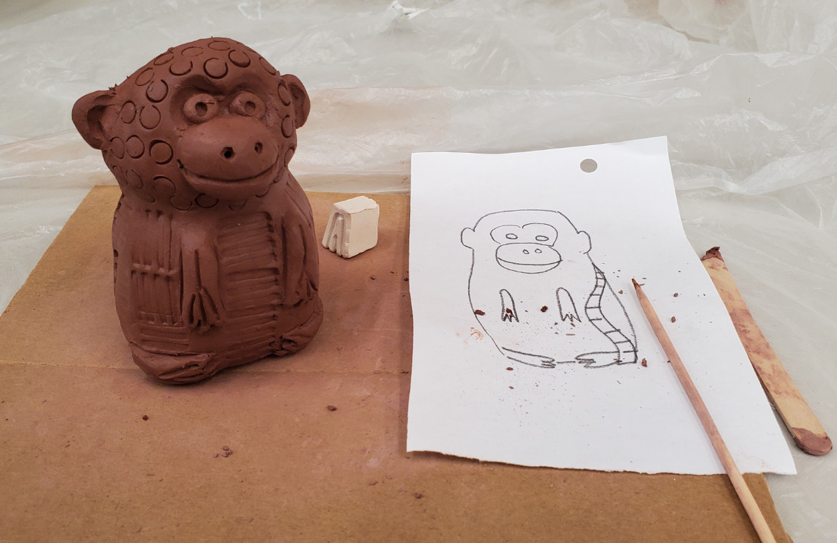 Sculpture d’argile texturée représentant un animal hybride inspiré du singe et de la souris. La sculpture est sur un carton avec des outils et un croquis.