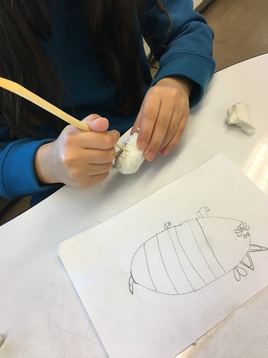 Mains d’enfant qui sculpte de l’argile polymère avec un outil de bois. Devant l’enfant se trouve un croquis d’animal hybride.
